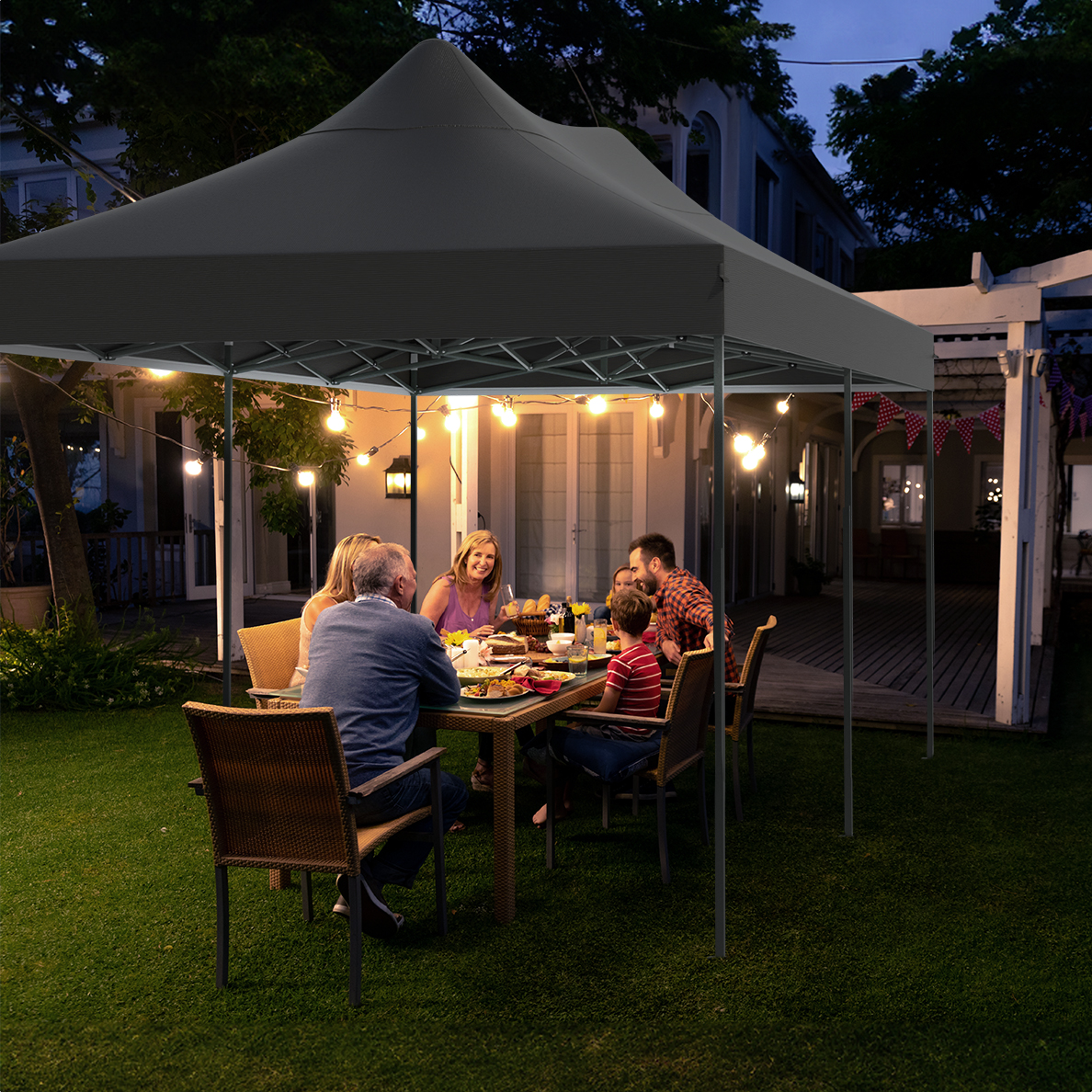 Family dining under a black tent at dusk. Table set with food, string lights, and a house in the background.