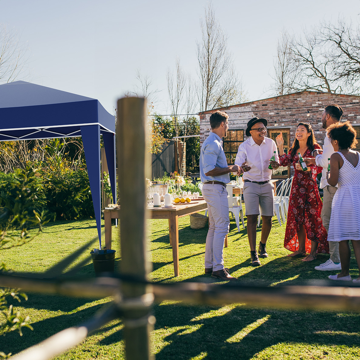 People in a garden, gathered around a table. A blue gazebo is on the left. Sunny, outdoor setting.