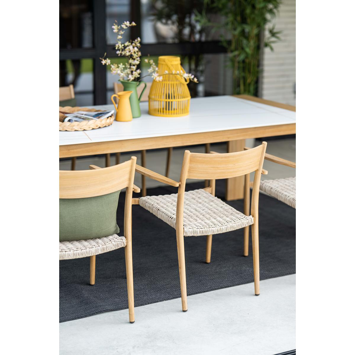 Outdoor dining scene: wooden chairs with woven seats around a table, and greenery.