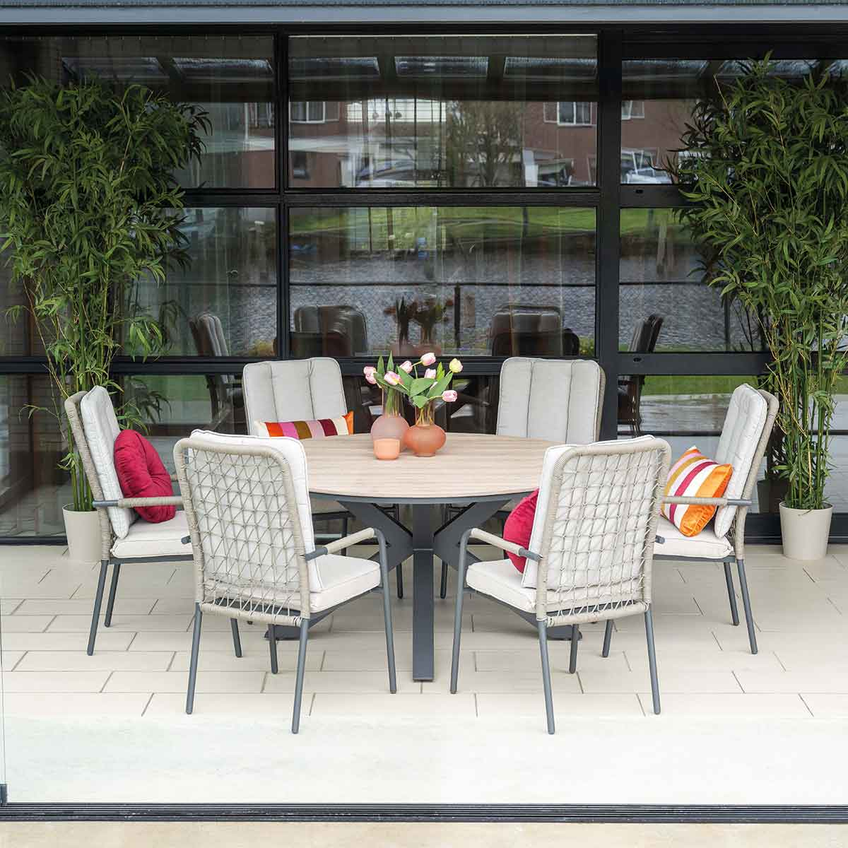 Outdoor dining set with a round table and chairs. Red pillows on the chairs. Green plants and glass windows.