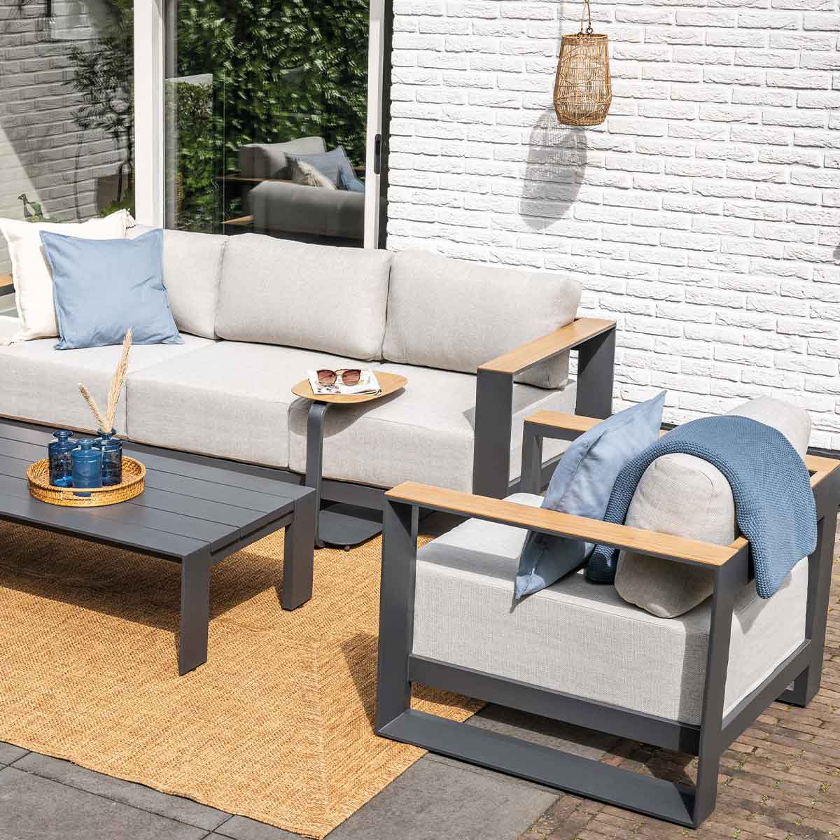 Mobilier de jardin : canapé, fauteuil, table basse sur un tapis tissé, avec coussins. En arrière-plan, un mur de briques blanches.