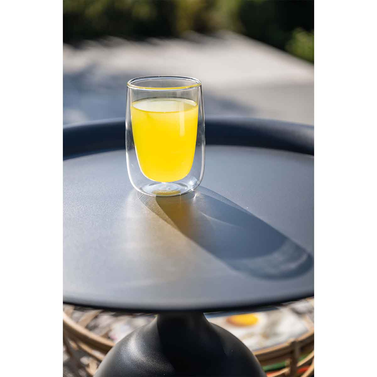 Un verre à double paroi avec liquide jaune est posé sur une table noire. Le fond montre la lumière du soleil et des arbres verts.