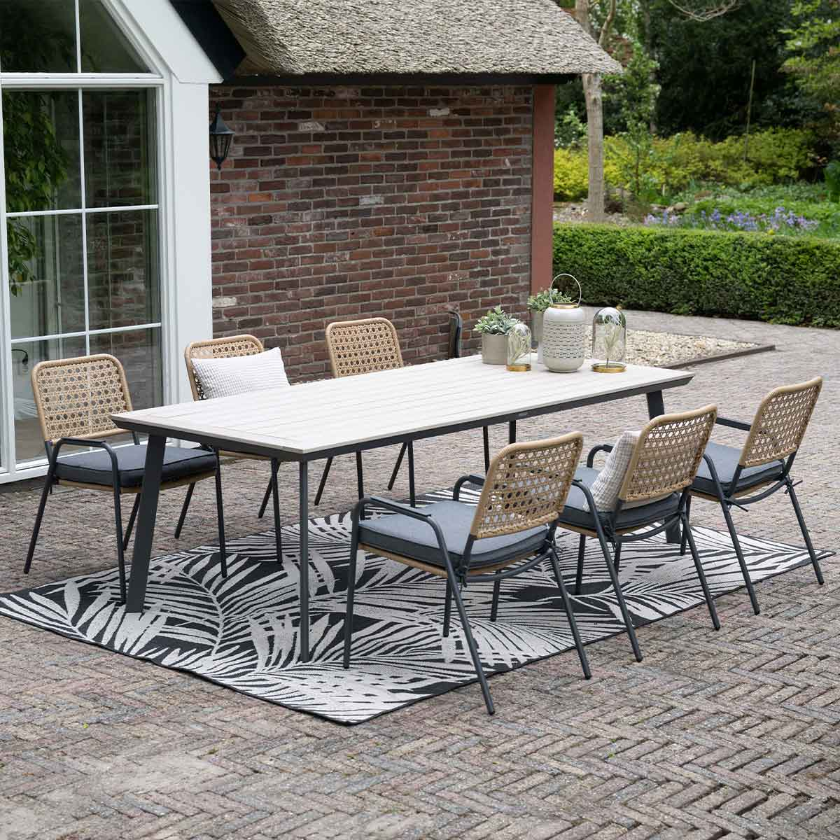 Outdoor dining set. Table, six chairs, patterned rug. Background: brick wall, greenery, blue sky, and a thatched roof.