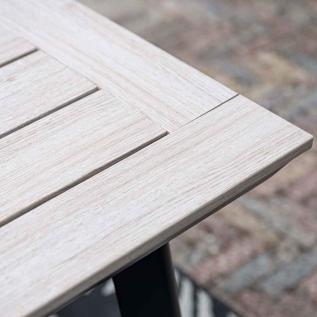 Close-up of a light-colored wooden table. Shows the corner of the table with wood grain detail. Black leg is visible.