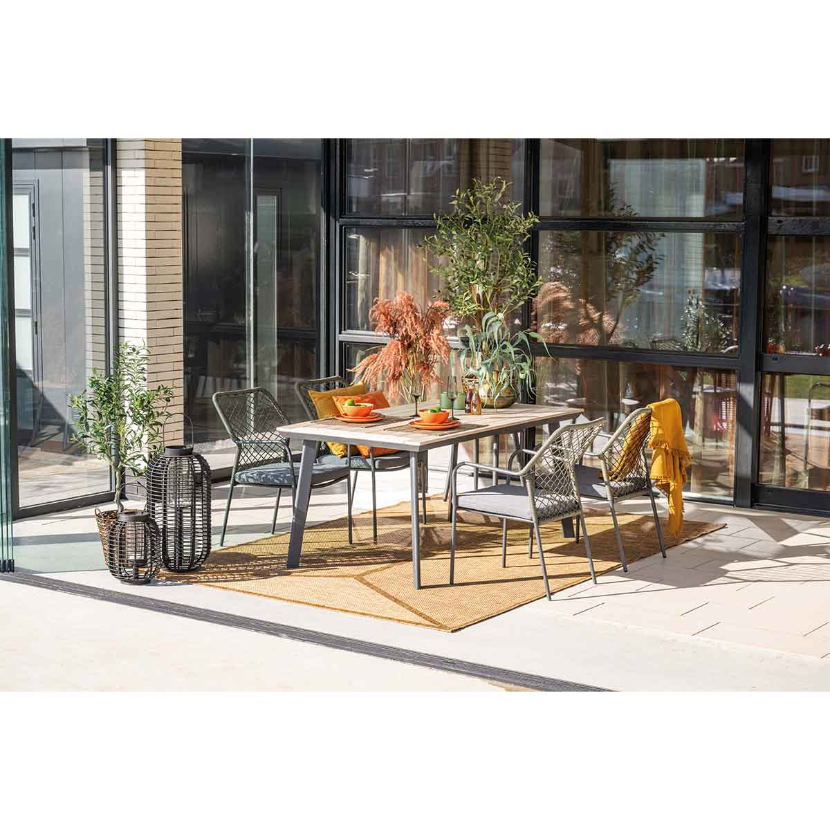 Outdoor dining setting with table, chairs, decor. Background: glass doors, plants, rug. A yellow blanket on a chair.