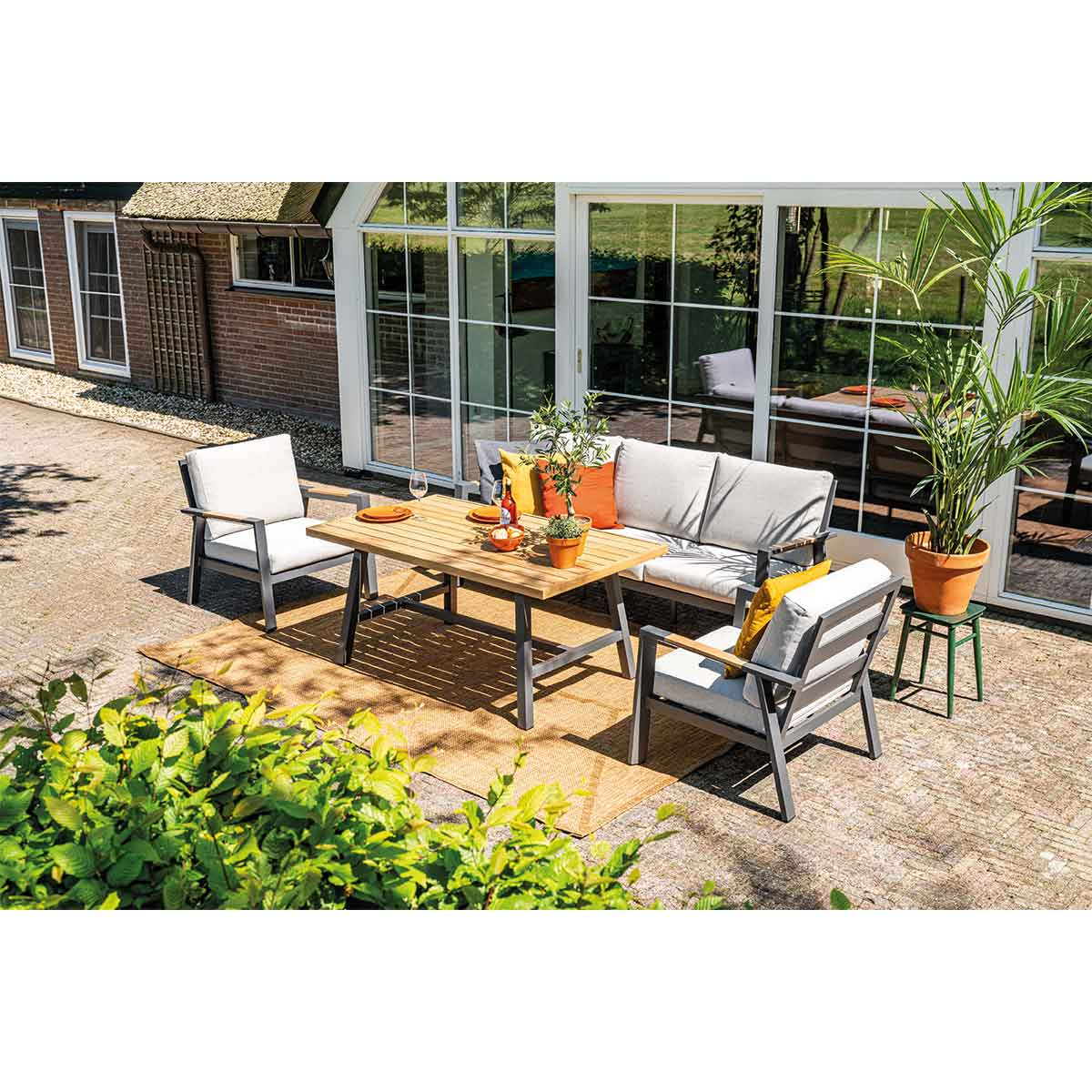 Terras met tafel, stoelen, bank en planten. Tafel met eten, oranje kussens en een tapijt.