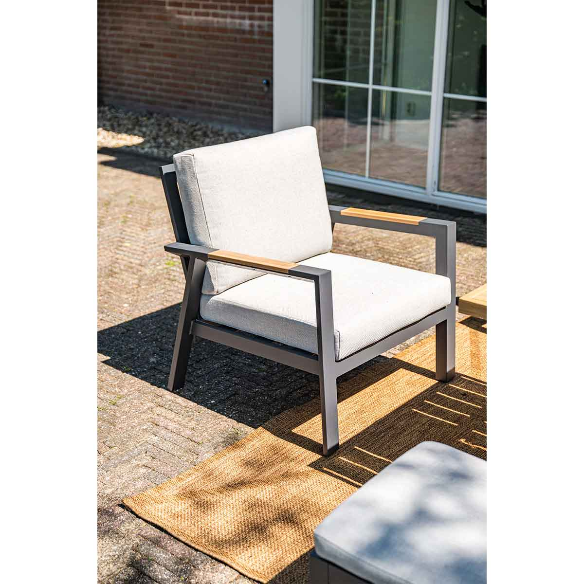 Grijze fauteuil met licht kussen, houten armleuningen, op een kleed en een bakstenen achtergrond.