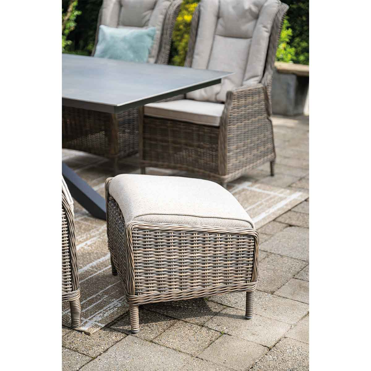 Wicker footstool with a light cushion, on a patio with a table and chair in the background.