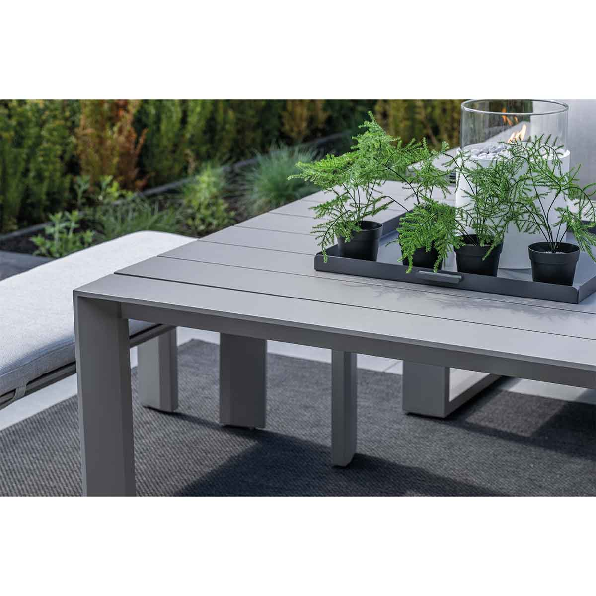 Table grise avec plantes et récipient en verre avec feu, banc et verdure en arrière-plan.