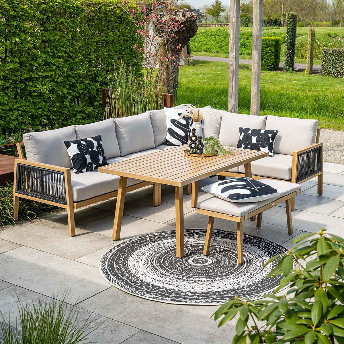 Een houten tafel, een bank en een bank op een terras, omringd door groen, met een rond tapijt.