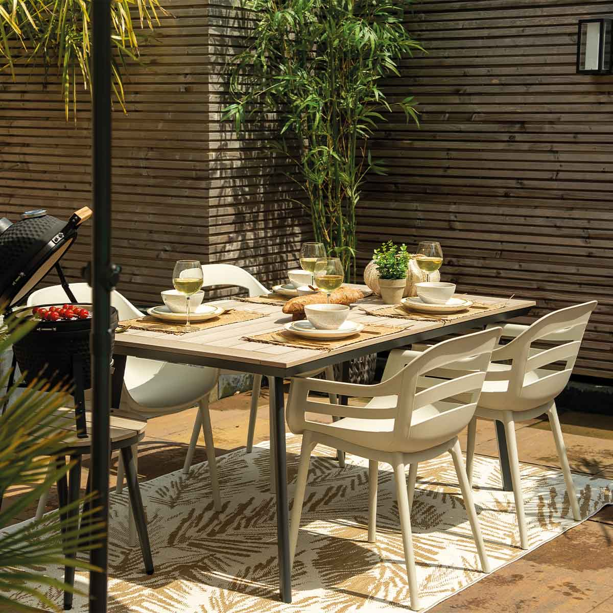 Outdoor dining set with table, chairs, food, and grill. The background features bamboo and wooden fence.
