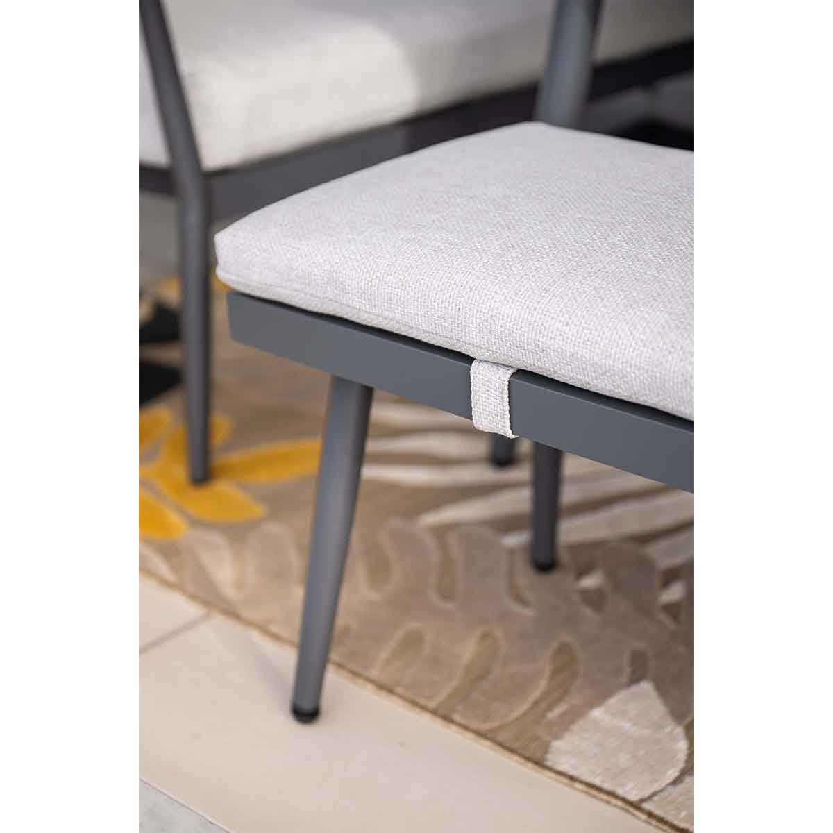 Close-up of bench with gray cushion. Gray legs, rug with leaf pattern, and tile floor.