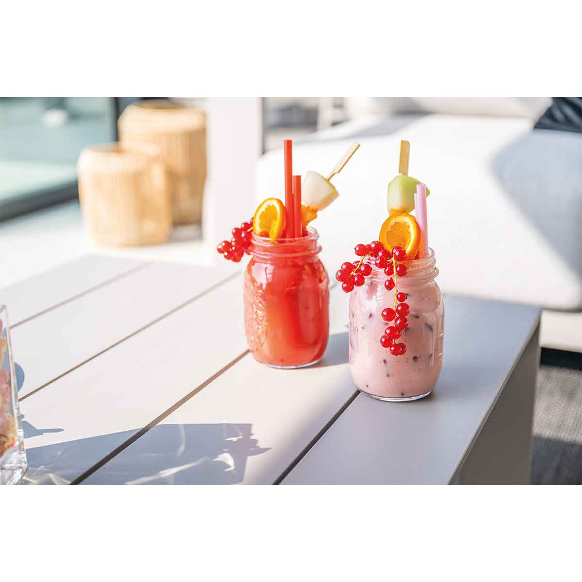 Twee weckpotten met drankjes en fruitgarnituren staan ​​buiten op een witte tafel.