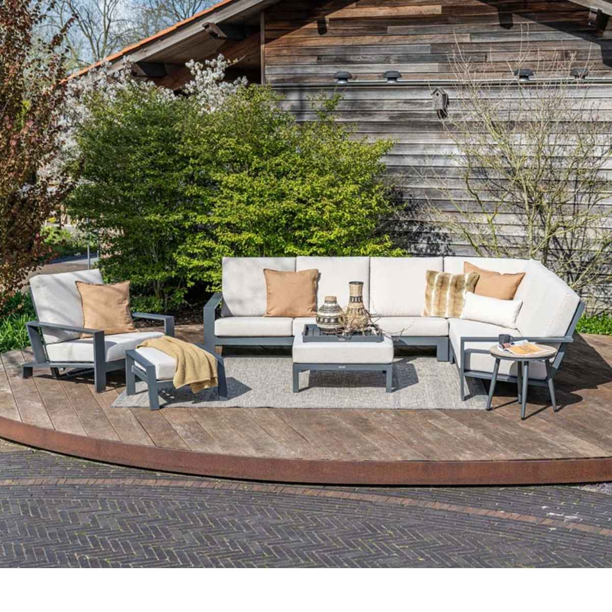 Outdoor sectional sofa with pillows, side table, and rug on a wooden deck with background.