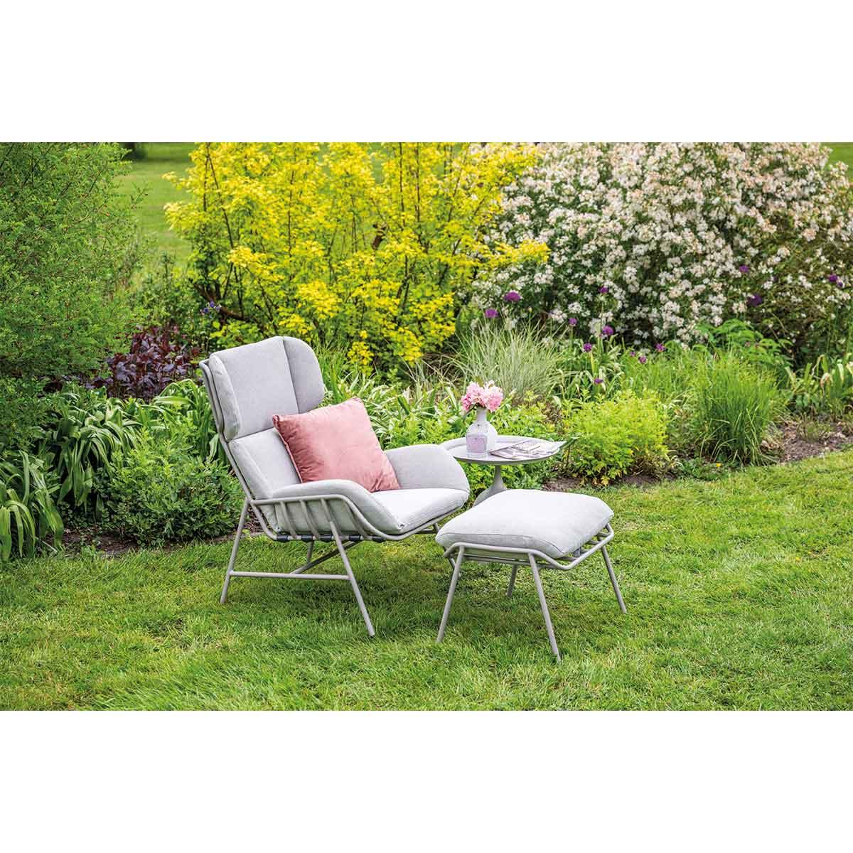 A gray chair and ottoman set on a grassy lawn. Pink pillow, flowers and plants surround the set.