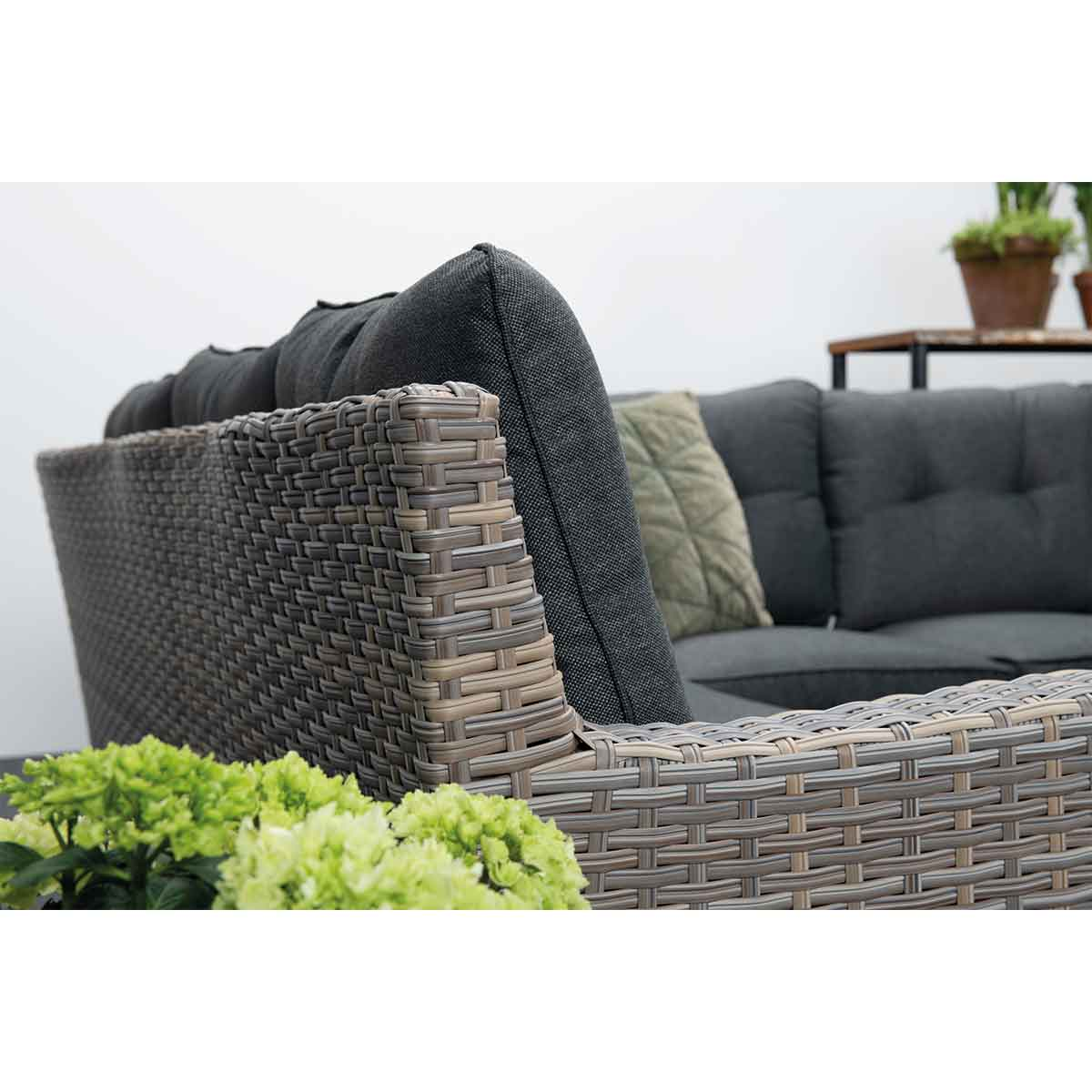 Close-up van grijze wicker bank met donkergrijze kussens. Groene planten en witte achtergrond.