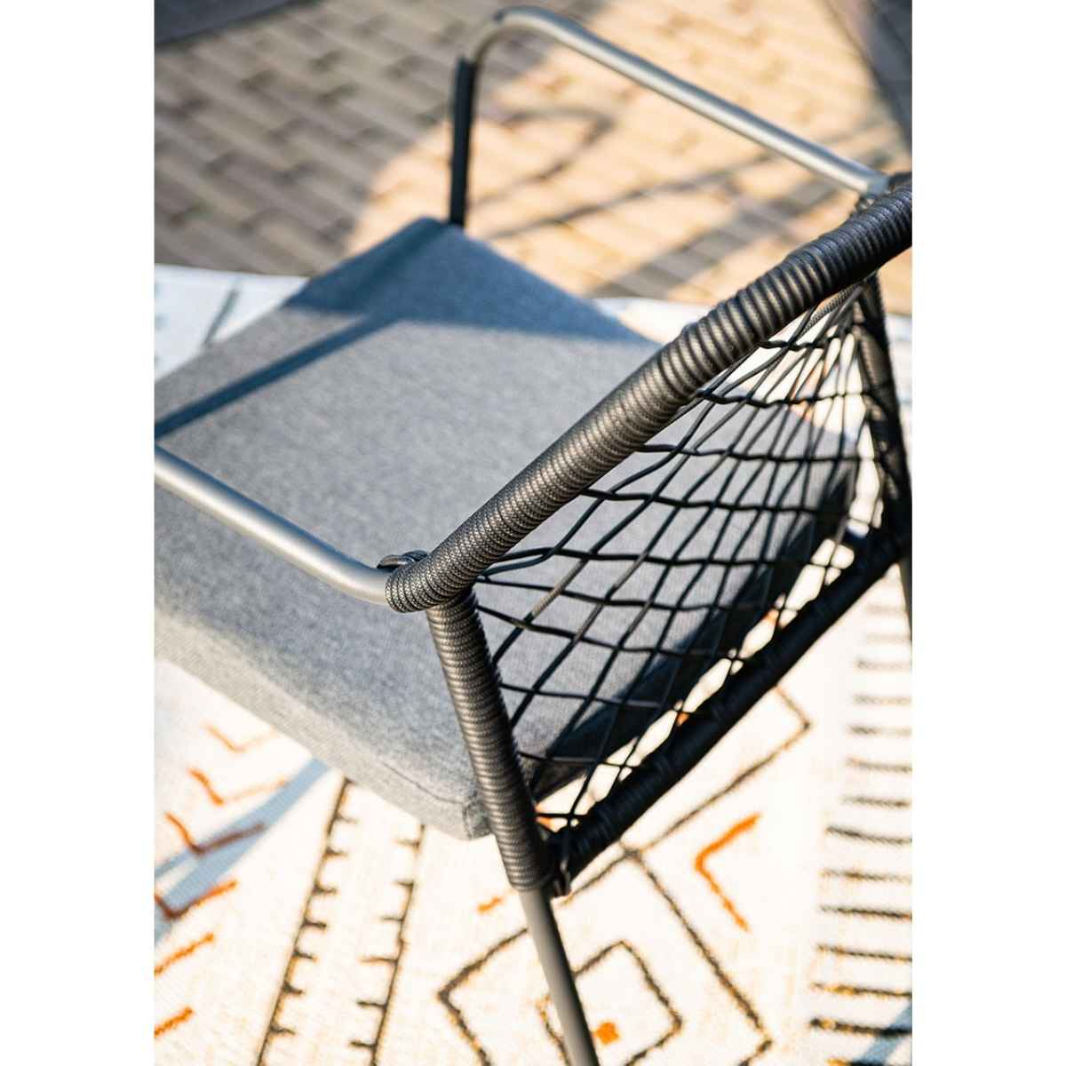 Chaise noire et grise avec un dossier tissé et un coussin gris, posée sur un tapis à motifs.