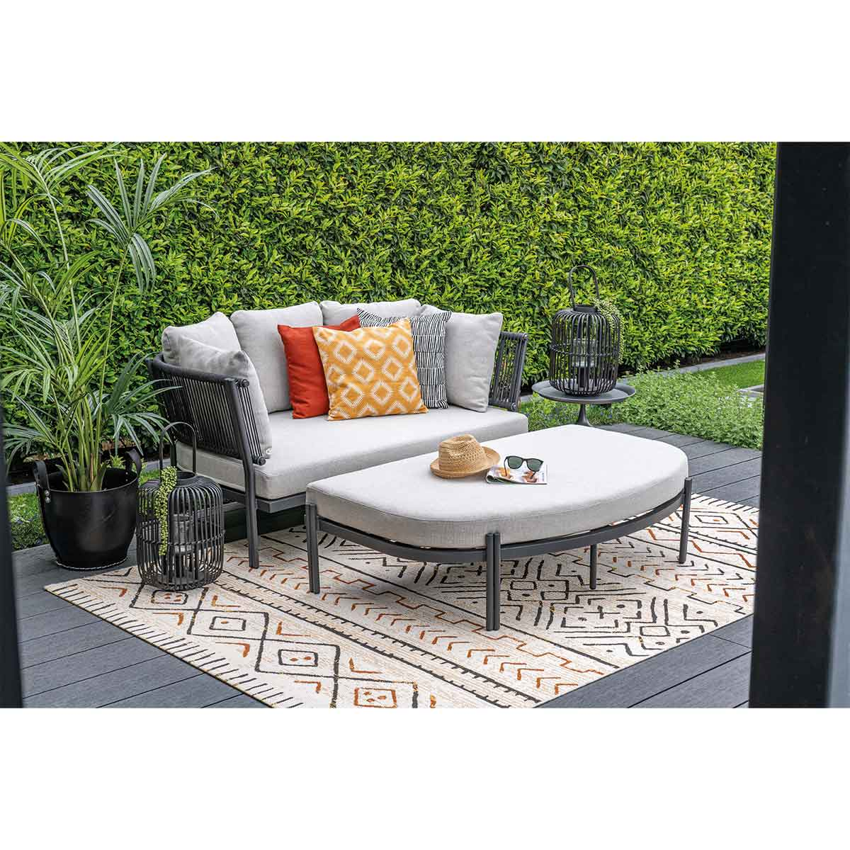 Outdoor scene with sofa, ottoman, rug, plants, and accessories, on a wooden deck.