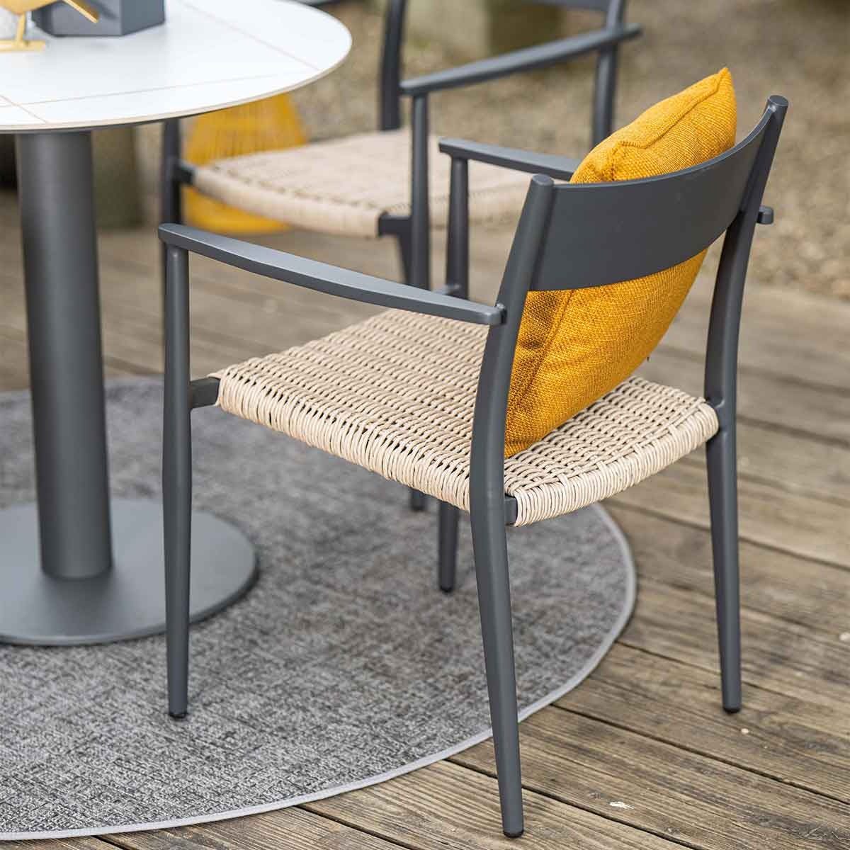 Grijze stoel met geweven zitting en geel kussen. Naast een kleine ronde tafel.