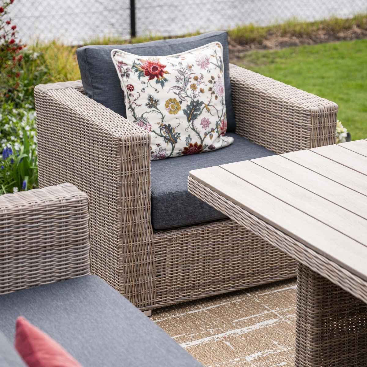 Chaise en osier avec coussin floral et coussin gris à côté d'une table en bois.