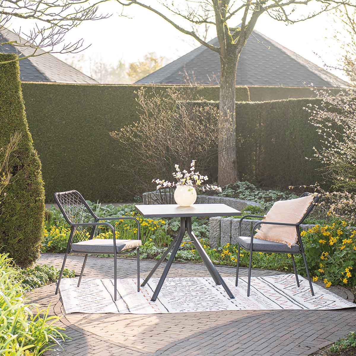 Outdoor scene with table, two chairs, flowers, and greenery. Table is black, chairs are gray.