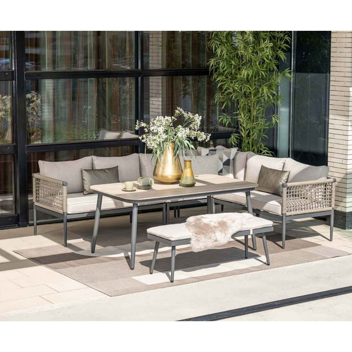 Salon de jardin : table, banc, deux canapés et tapis. Coussins beiges, cadres gris. Fleurs blanches dans un vase.