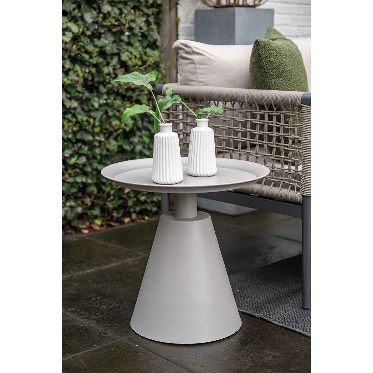 Een grijze bijzettafel buiten met twee witte vazen met groene planten. Een stoel op de achtergrond.