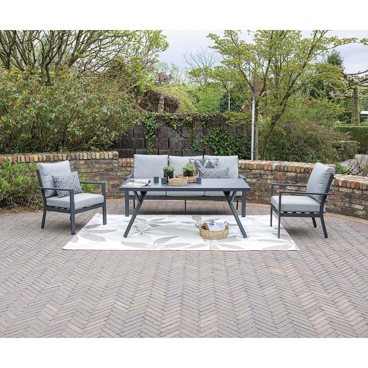 Outdoor patio set with grey furniture and a brick wall backdrop. Green plants and trees in the background.