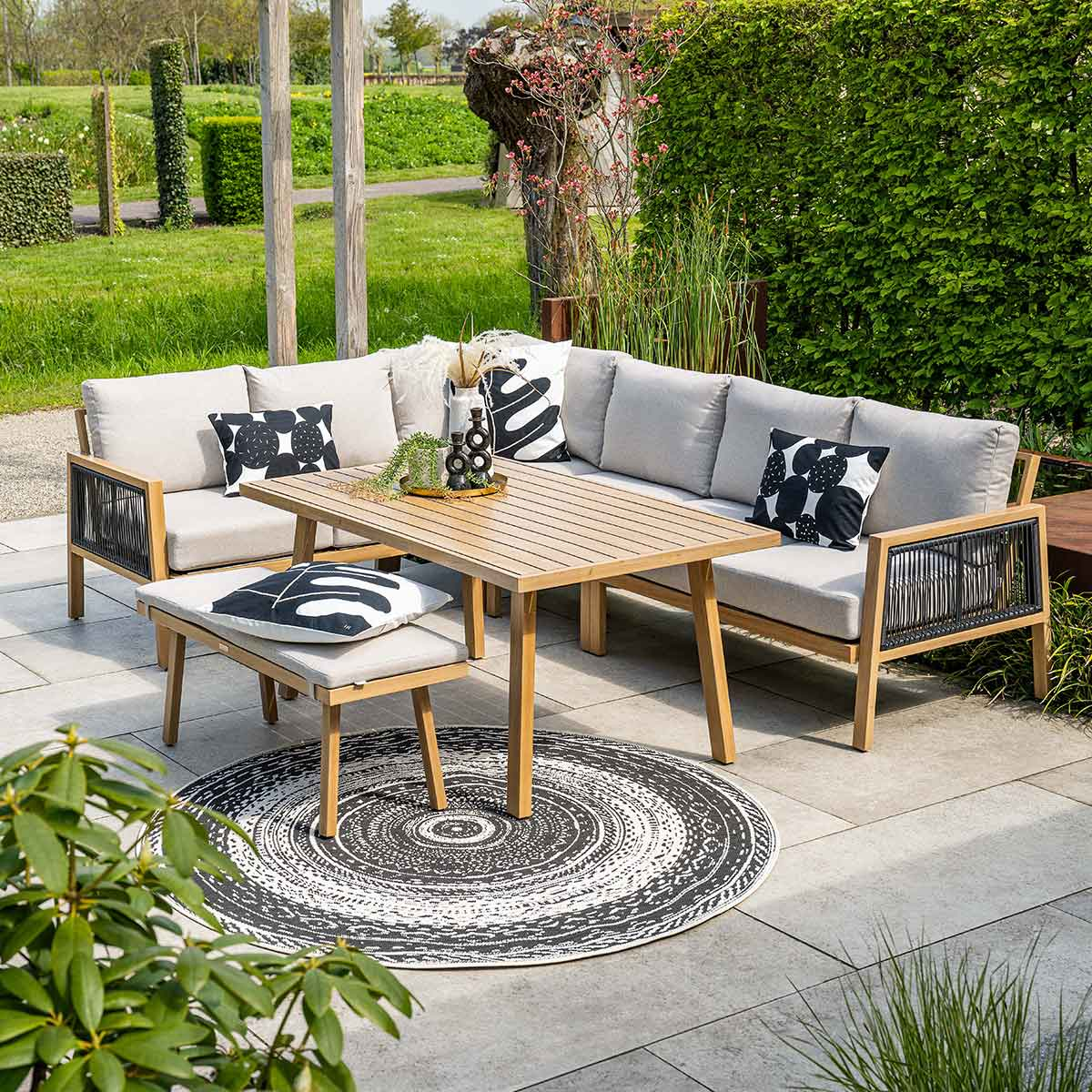 Buitenzitgroep: lichte houten tafel met zitplaatsen, kussens, vloerkleed en planten.
