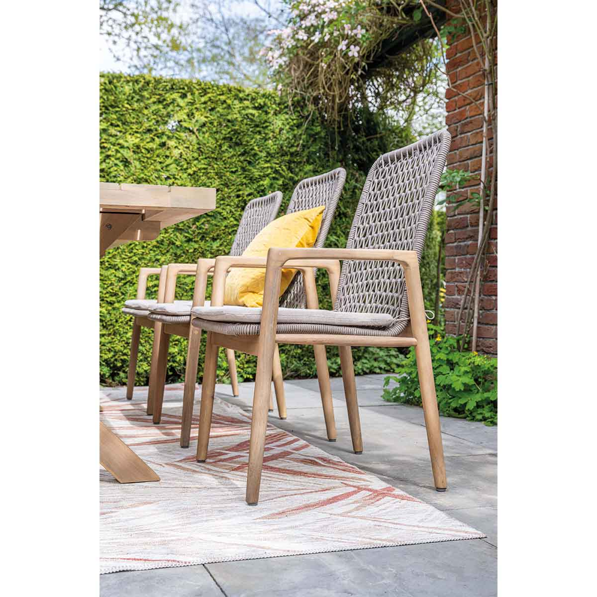 Three outdoor chairs around a table, with a garden background. One has a yellow pillow. Sunny light.