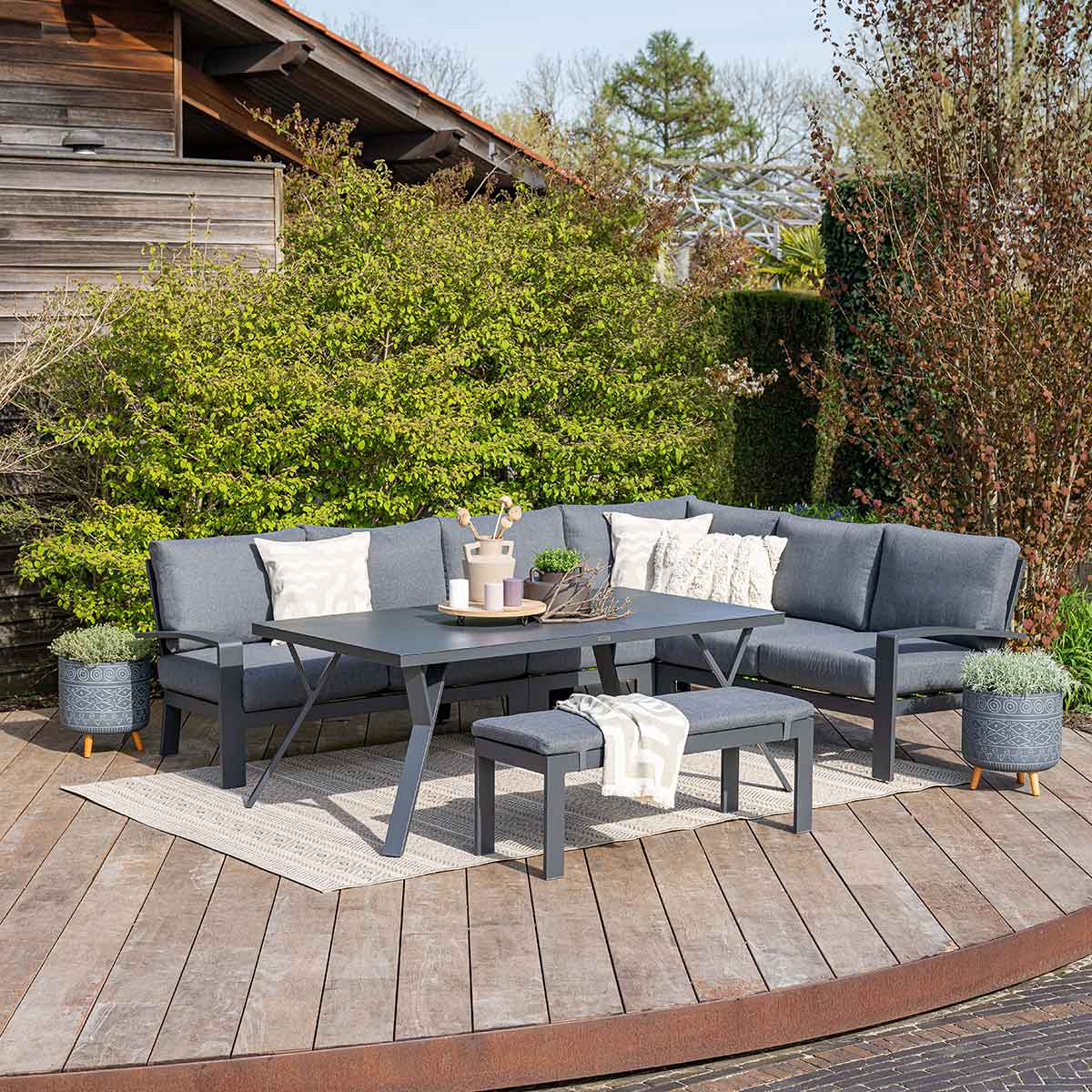 Buitenmeubelset: grijze hoekbank, tafel, bankje en twee potplanten, op een houten terras.