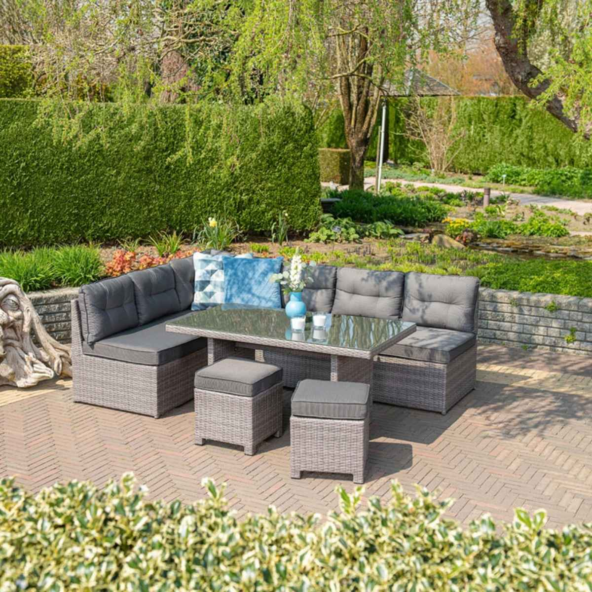 Canapé d'angle gris extérieur avec table, poufs et fond de jardin. La scène est en plein jour.