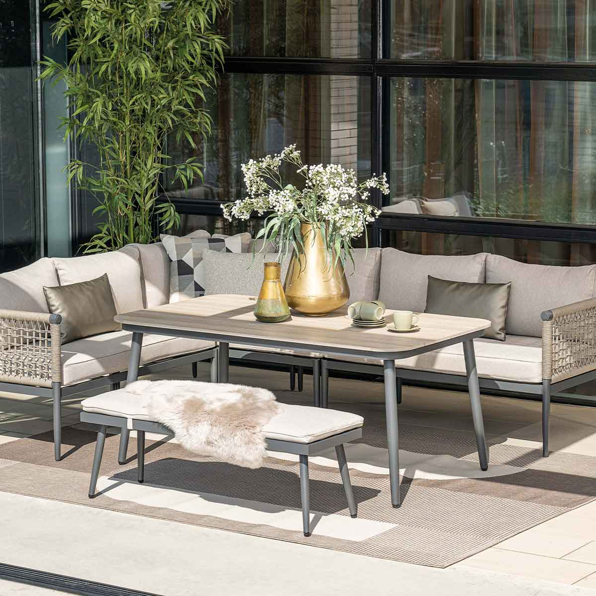 Patio furniture set: table, bench, sofa and chairs. Beige cushions, grey frames. Rug, vase, and flowers.
