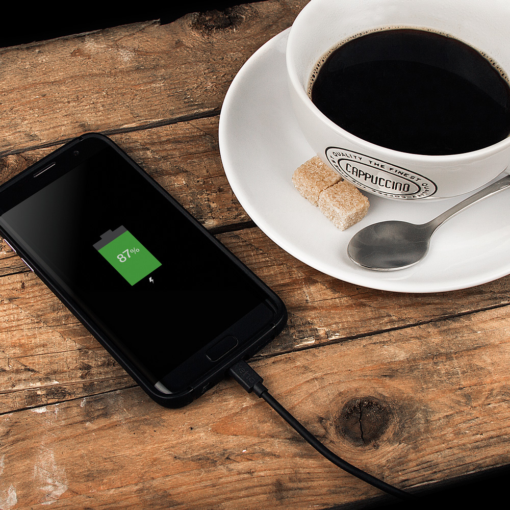 Ein schwarzes Smartphone, das auf einem Holztisch geladen wird. Eine Tasse Kaffee und Würfelzucker sind in der Nähe.