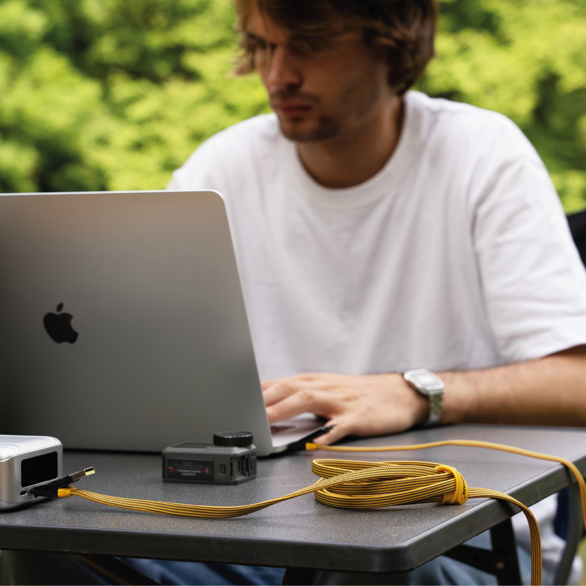Mann, der im Freien einen Laptop benutzt. Ein Kabel ist angeschlossen. Eine Powerbank und eine Kamera liegen auf dem Tisch.