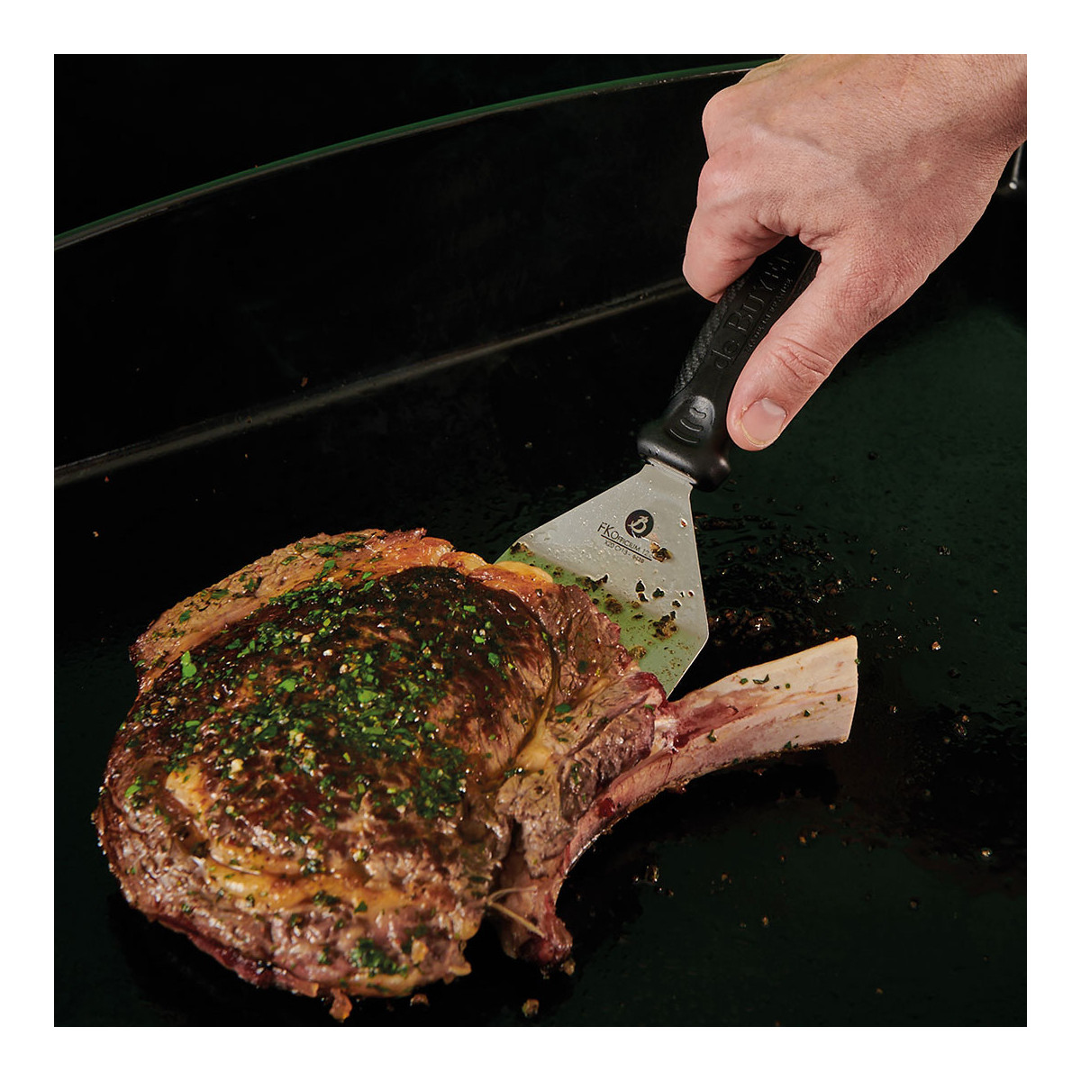 Eine Hand, die mit einem Pfannenwender ein gegrilltes Steak auf einer schwarzen Oberfläche wendet.