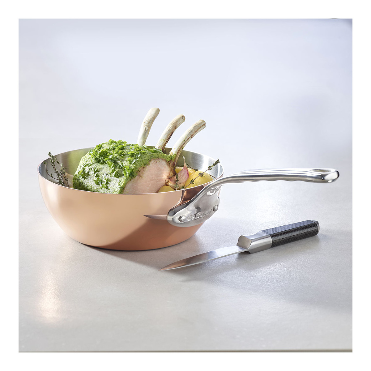 Kupfer-Kochtopf mit Griff, enthält Fleisch mit Kräutern und ein Messer auf weißer Oberfläche.