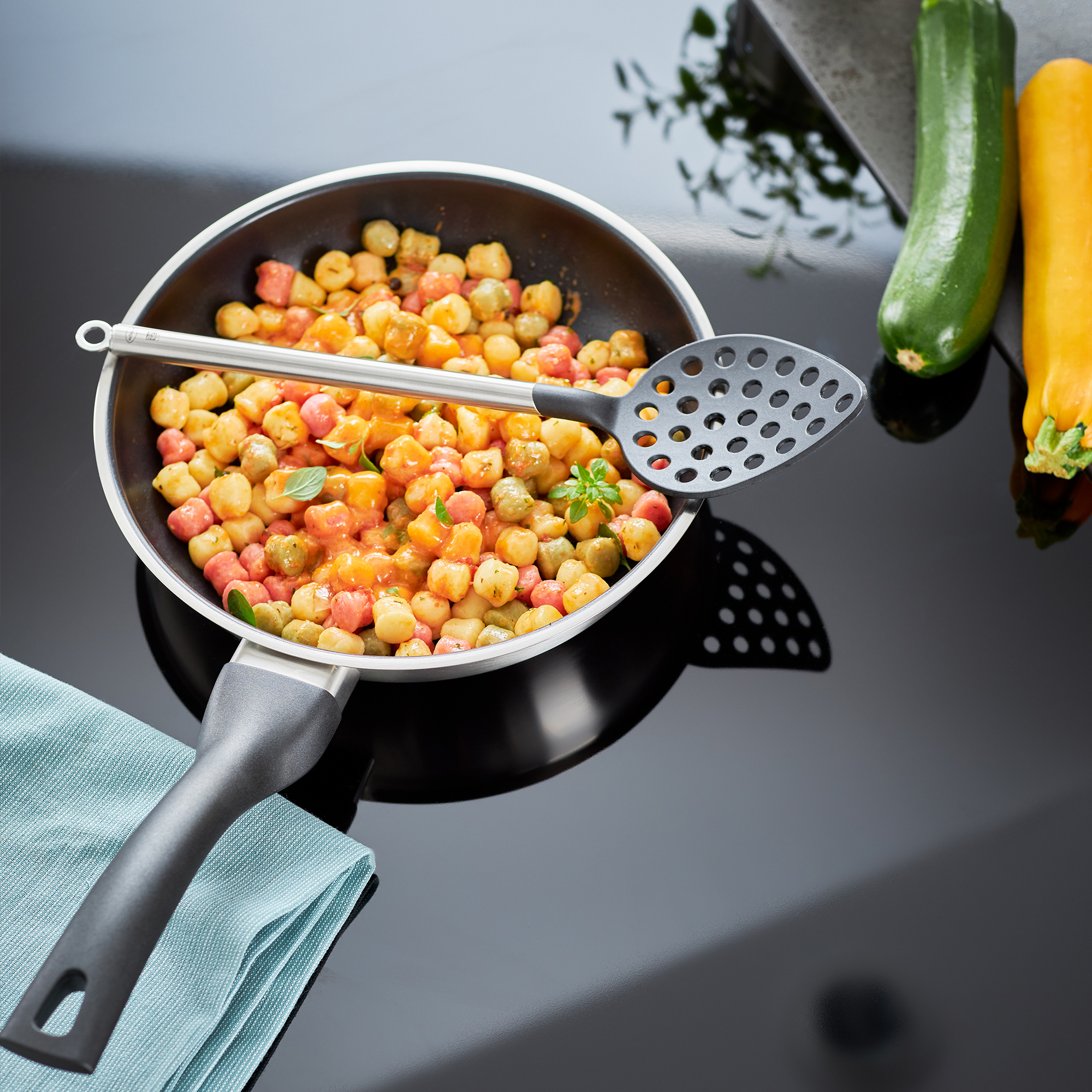 Pfanne mit Gnocchi in Sauce. Ein Schlitzlöffel liegt in der Pfanne. Zucchini im Hintergrund.