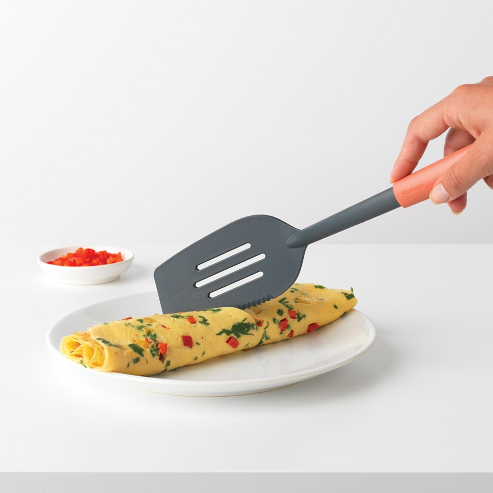 Ein Spatel hebt ein Omelett auf einem weißen Teller an, mit roten Paprika.