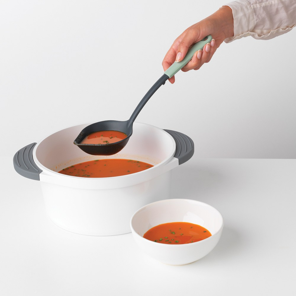 Hand schöpft Suppe mit grünem Griff aus Topf in Schüssel.
