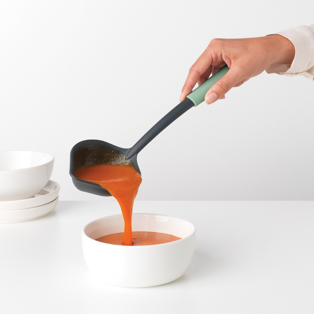 Eine Person gießt orangefarbene Suppe aus einer schwarzen Kelle in eine weiße Schüssel.