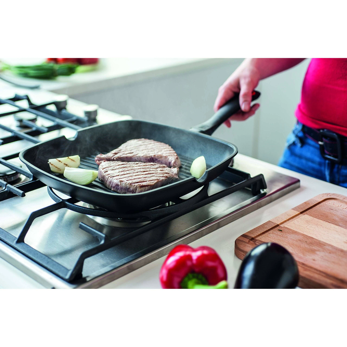 Steaks werden auf einer Grillpfanne auf dem Herd gebraten. Eine Zwiebel und Paprika sind in der Nähe.