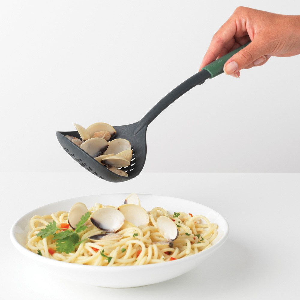 Una persona tiene un cucchiaio forato nero e verde pieno di vongole, sopra la pasta in una ciotola bianca.