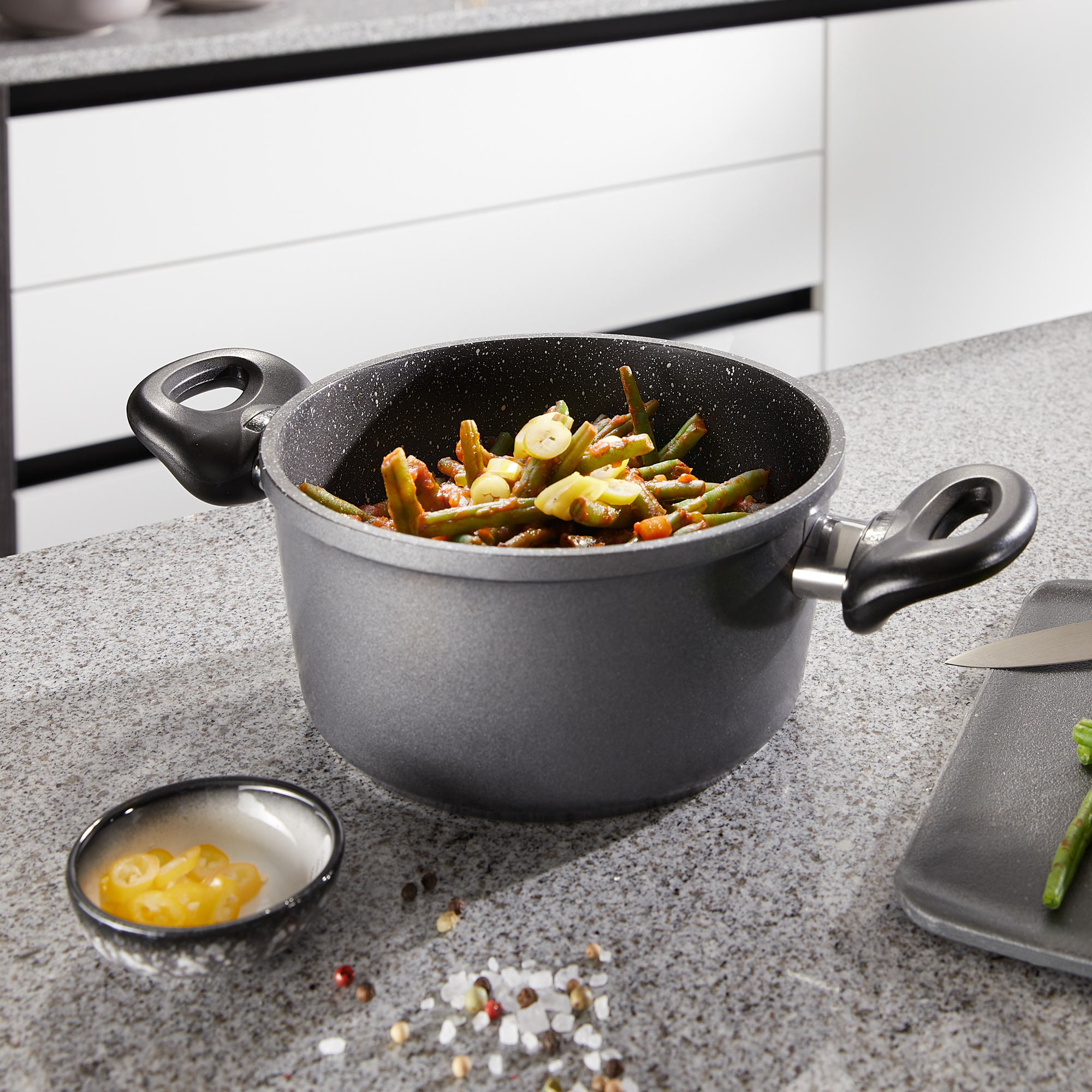 Garnek z warzywami w środku. Znajduje się na szarym blacie z małą miską i nożem.