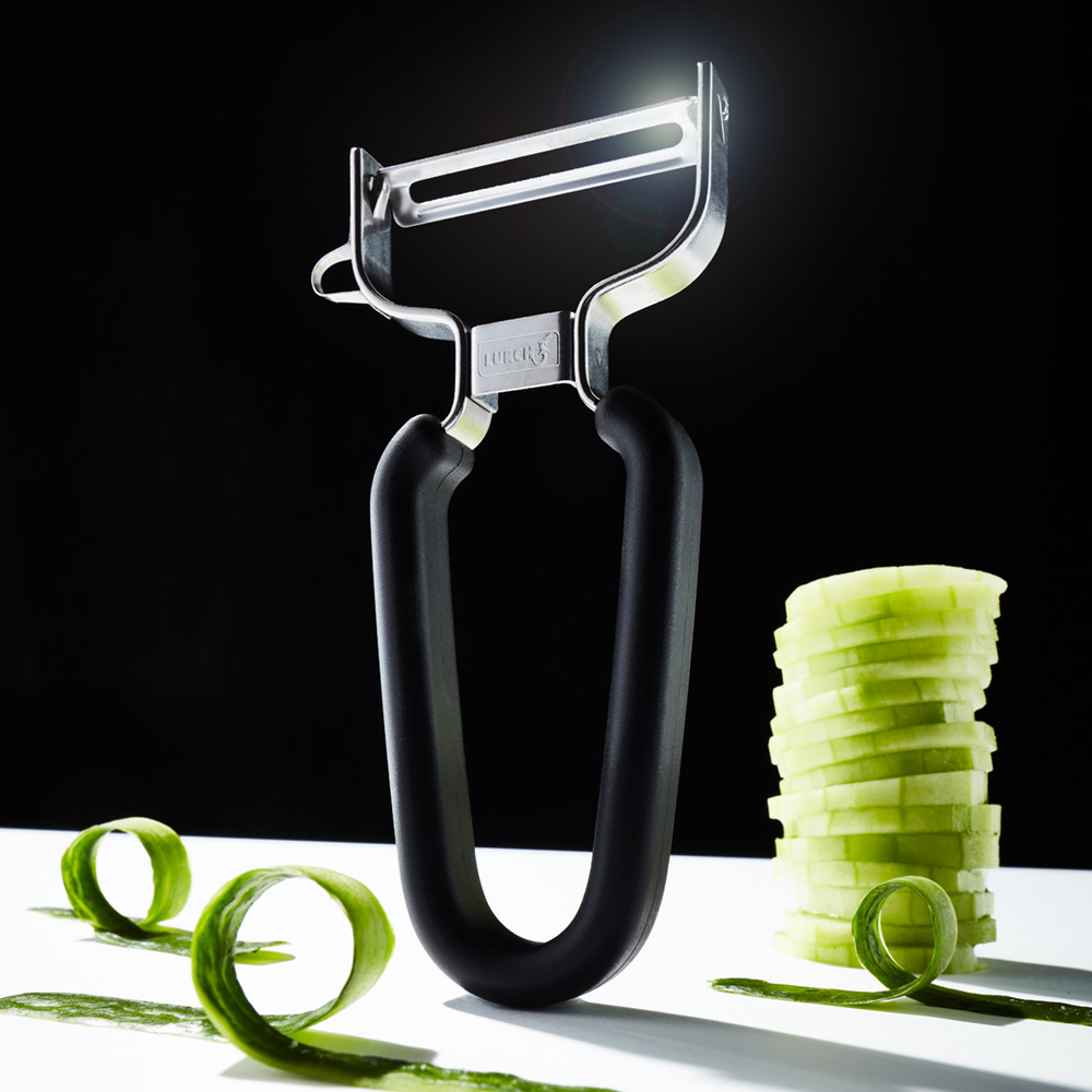 Gemüseschäler mit Gurkenscheiben und Locken auf weißer Oberfläche, dunkler Hintergrund.