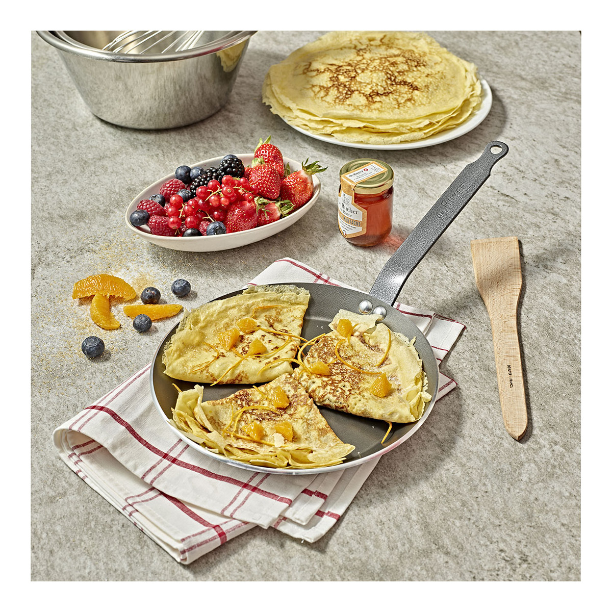 Pancakes with orange slices and berries, a wooden spatula, and ingredients.