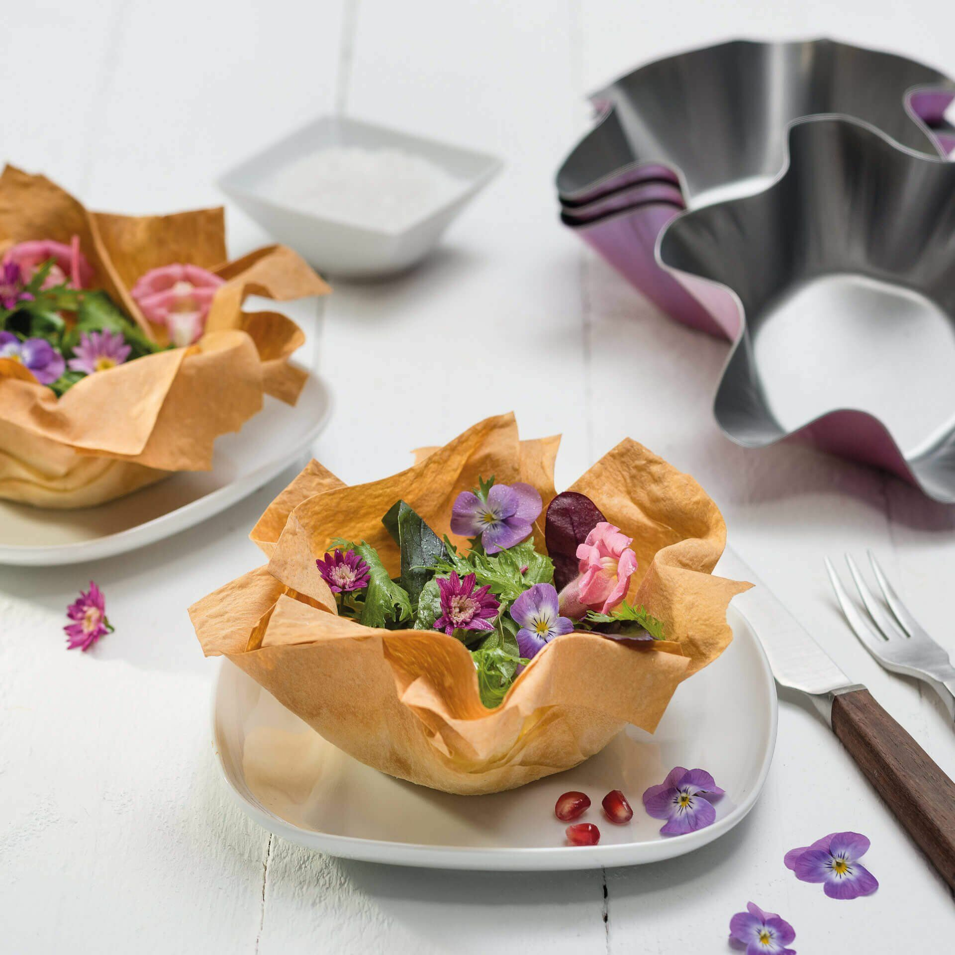 Zwei essbare Schalen mit Salat und Blumen, Messer und Gabel. Weiße Oberfläche.