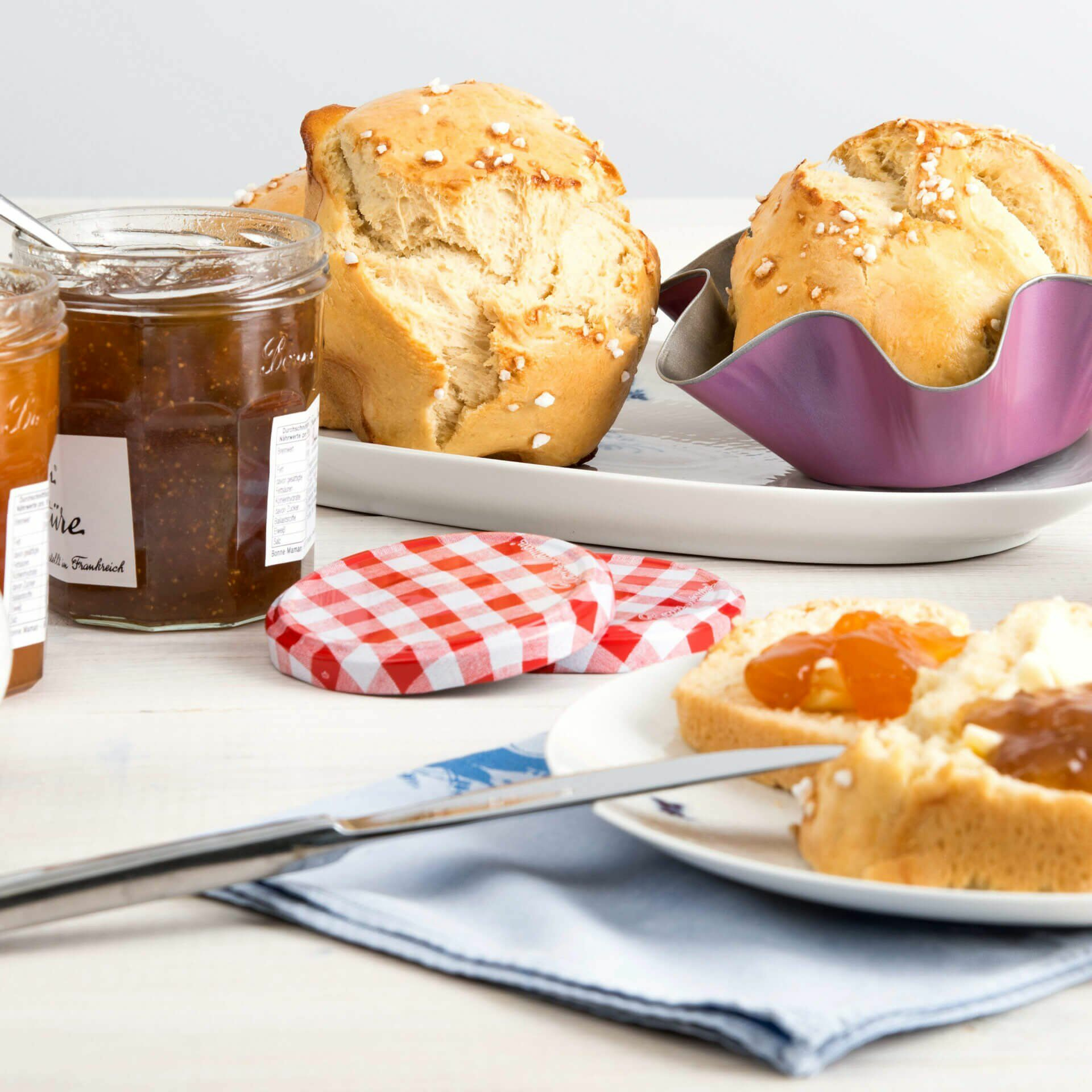Brot, Marmeladengläser und ein Messer auf weißer Oberfläche. Die Szene erinnert an ein Frühstück.