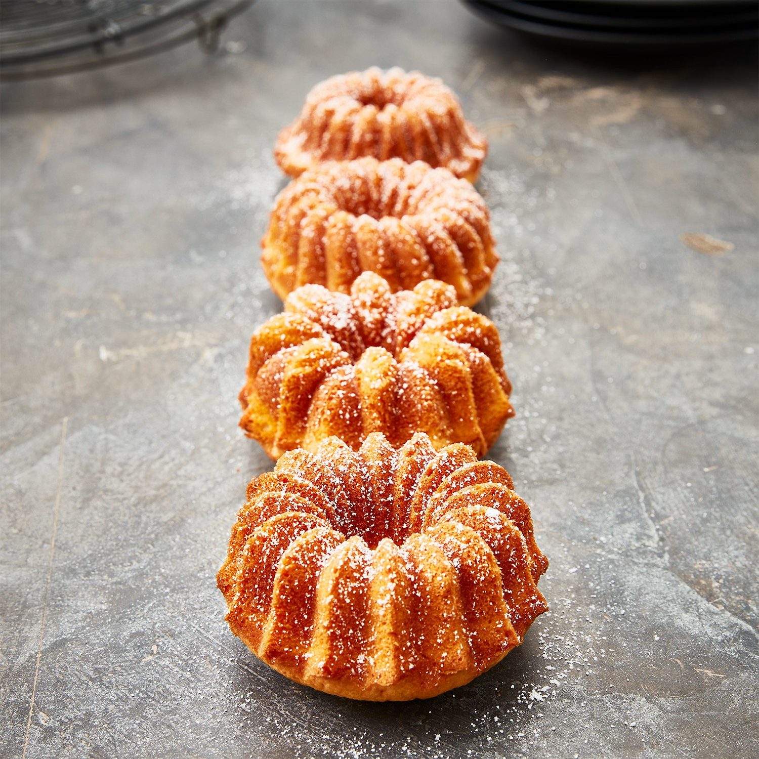 Vier kleine Gugelhupfkuchen, mit Puderzucker bestäubt, in einer Reihe angeordnet.