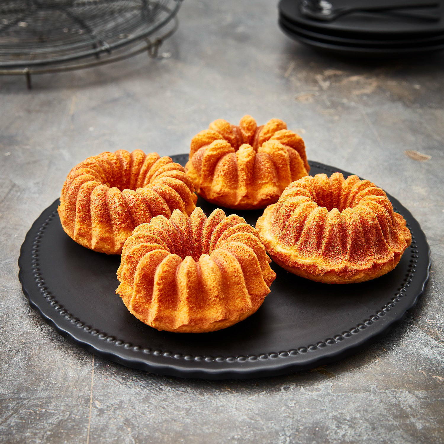 Vier goldbraune Mini-Gugelhupf-Kuchen auf einem schwarzen Teller auf grauer Oberfläche.